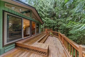 Beautiful large deck with built-in L-shaped bench attached to green house with lots of windows and forest in the background