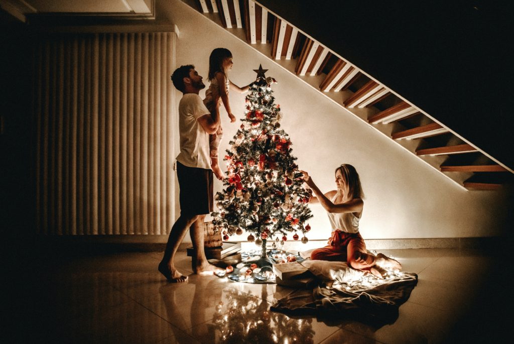 Dad holding young daughter to put star on top of Christmas tree with mom sitting on floor under stairs