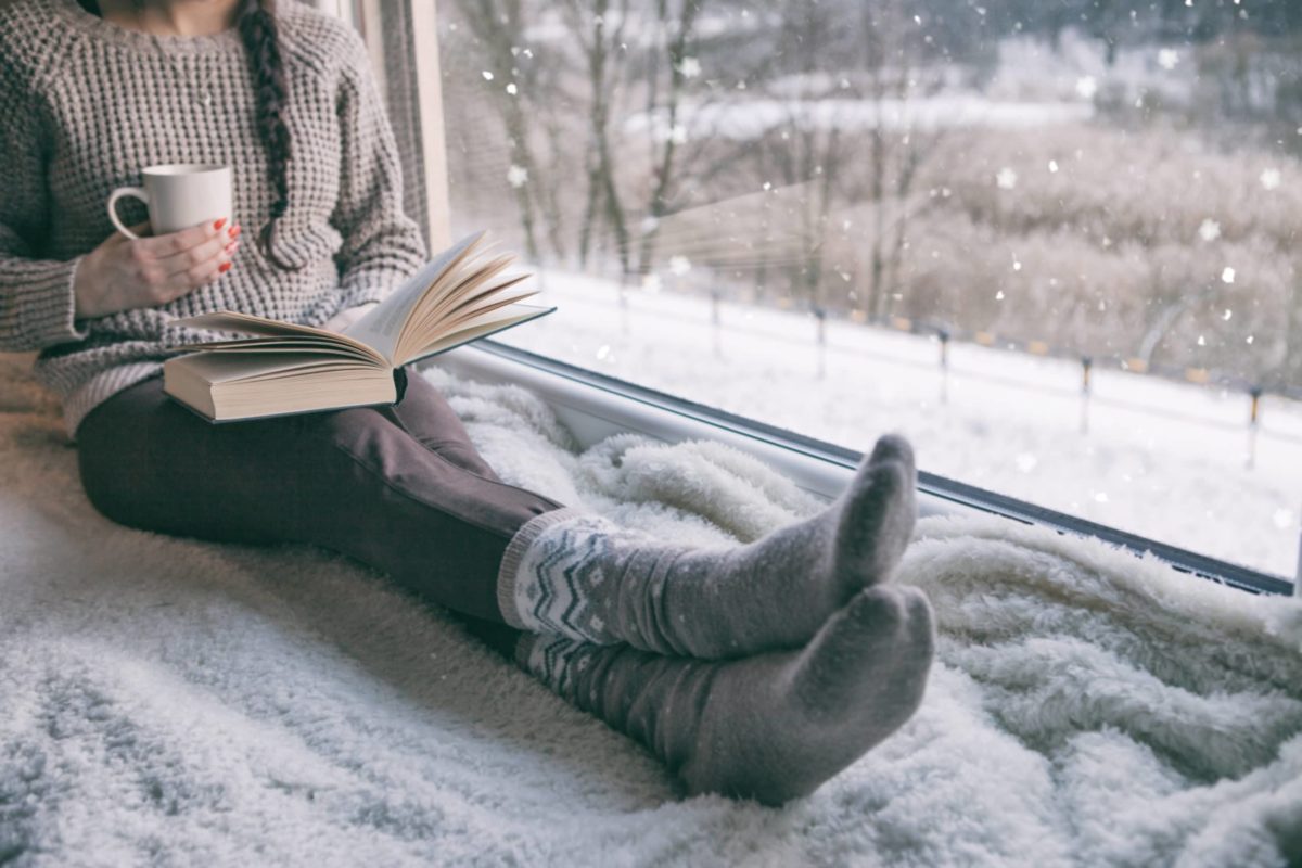 Woman in sweater, leggings, and cozy socks reading by a large window with snow outside